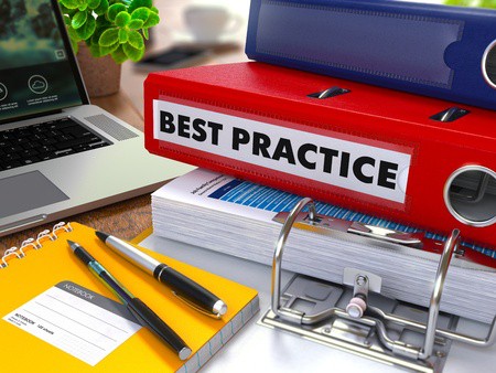 Red Ring Binder with Inscription Best Practice on Background of Working Table with Office Supplies, Laptop, Reports. 